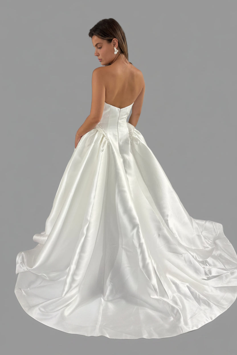 A woman modeling a white satin ball gown wedding dress from the back, showing a structured backless bodice and a full satin overskirt with a train, set against a background of other wedding dresses on racks.