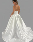A woman modeling a white satin ball gown wedding dress from the back, showing a structured backless bodice and a full satin overskirt with a train, set against a background of other wedding dresses on racks.