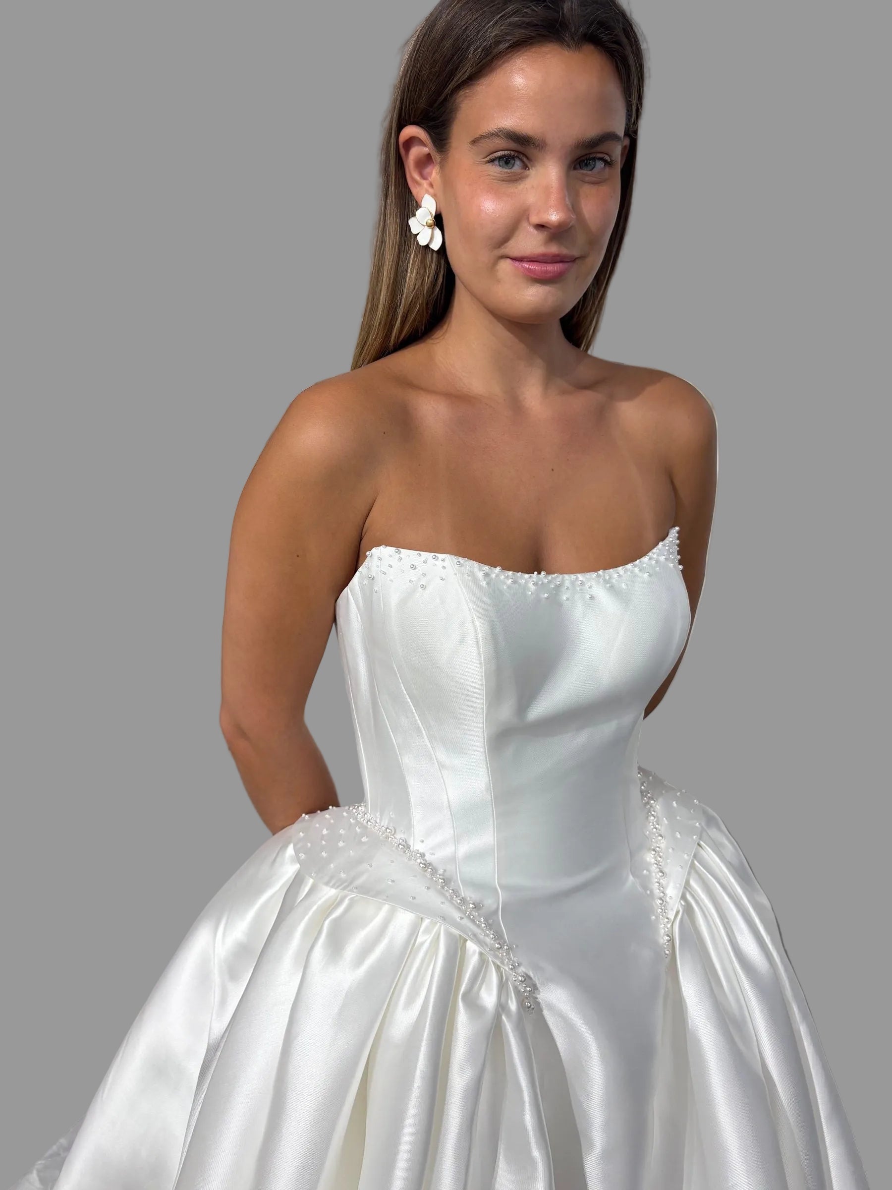 A woman wearing a strapless white satin ball gown with a full skirt, standing in a bridal shop with other white wedding dresses visible in the background.