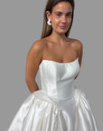 A woman wearing a strapless white satin ball gown with a full skirt, standing in a bridal shop with other white wedding dresses visible in the background.