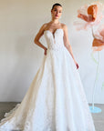 Woman in a white strapless lace wedding dress standing next to large floral arrangements.