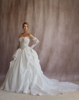 A woman modeling a strapless white wedding gown with long lace sleeves, a fitted lace bodice, and a voluminous satin overskirt with a train